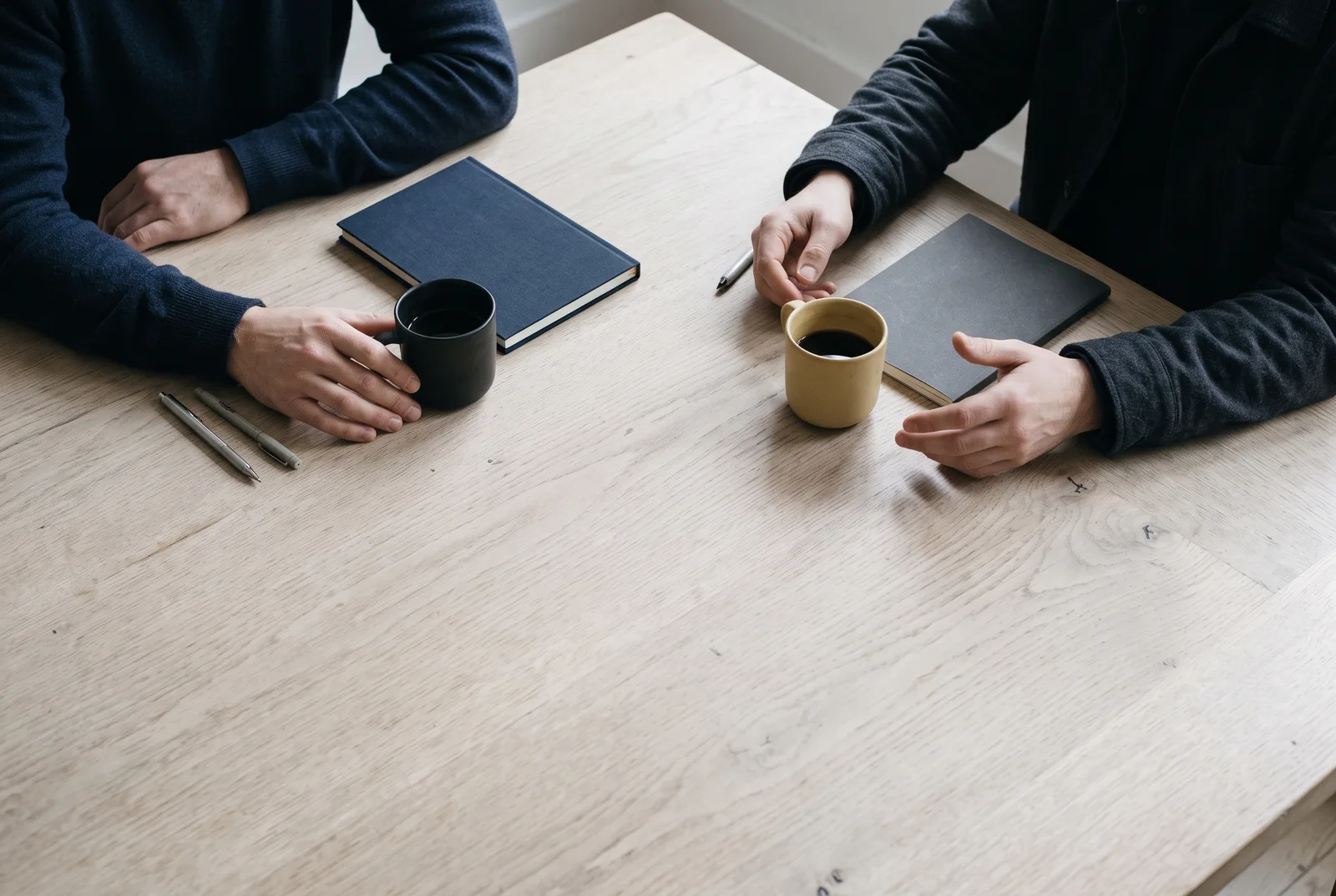 Two people’s hands and coffee cups across an oak table during a conversation