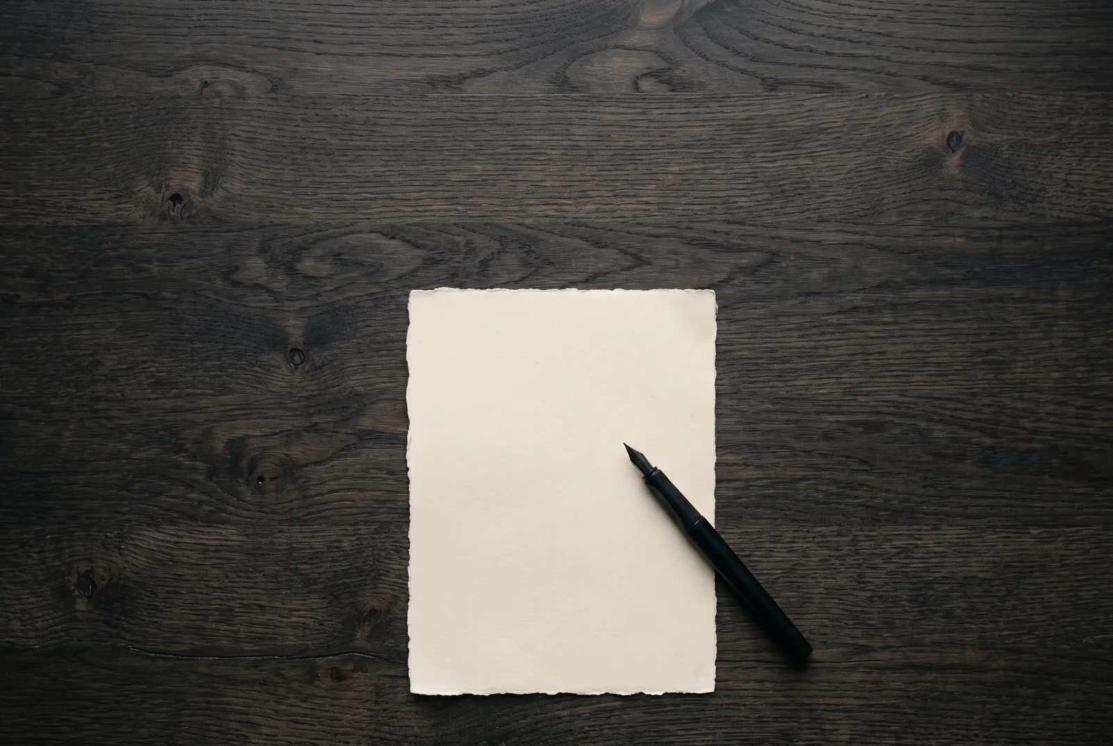 A blank sheet of cream paper and a fountain pen on a dark desk